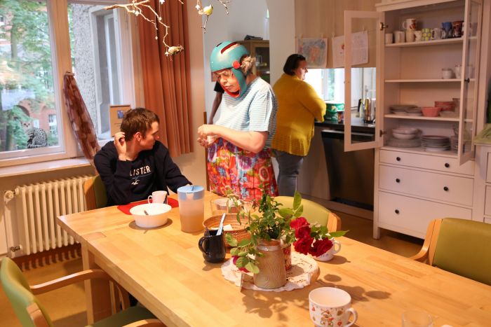 Wohnsituation in der Küche: eine Mitbewohnerin deckt den Tisch, eine andere spült ab, ein junger Mann istzt am gedeckten Tisch