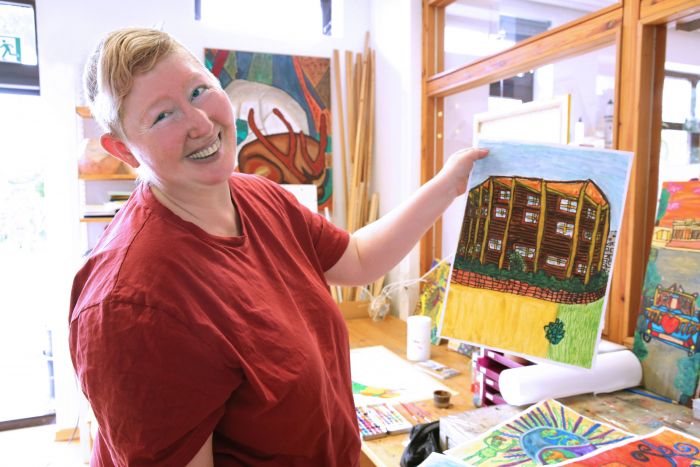 Eine glücklich lachende Malerin im roten Shirt präsentiert ihr Kunstwerk in die Kamera