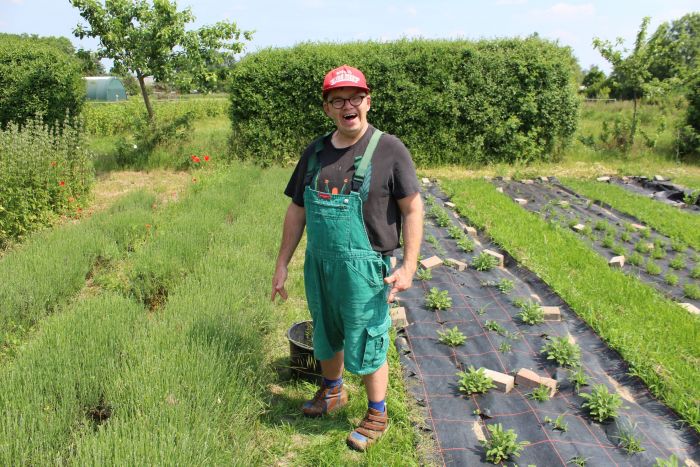Ein fröhlicher Mitarbeiter unserer Gärtnerei steht im Feld in grüner Latzhose und lächelt in die Kamera
