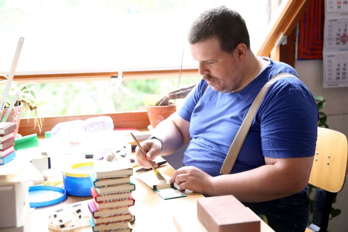 Ein junger Mann arbeitet sitzend in der Buchbinderei und bestreicht Buchrücken mit Leim