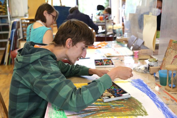 Ein junger Mann im kariertem Kapuzenpulli malt ein Bild mit Pastellfarben in unserer Kunstwerkstatt