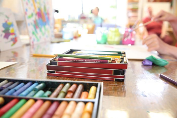 Ein Tisch mit Pastellfarben und bunten Stiften, im Hintergrund maldende Menschen