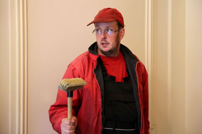 Ein junger Mitarbeiter von Problemlos in roter Kleidung und roter Kappe mit Besen in der Hand, zur Seite schauend