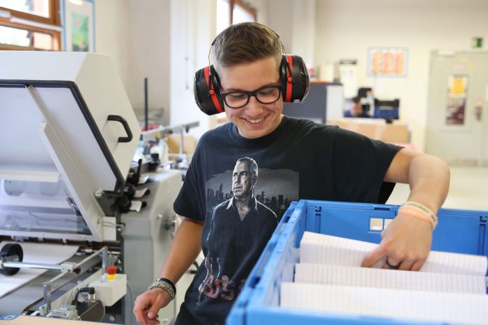 Ein junger Mann mit Kopfhörern arbeitet lächelnd an der Papiermaschine und nimmt einen Block mit Blättern aus einer blauen Kiste heraus