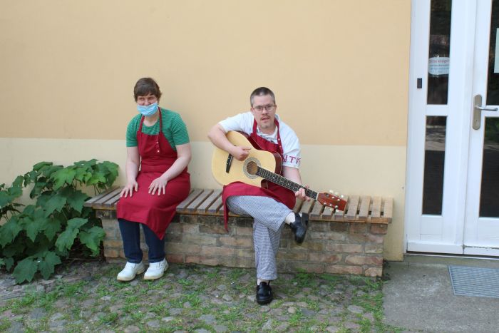 Zwei Mitarbeiter unserer Gärtnerei sitzen auf einer Bank vor dem Haus, beide mit roter Schürze, Frau lächelt und Mann spielt Gitarre